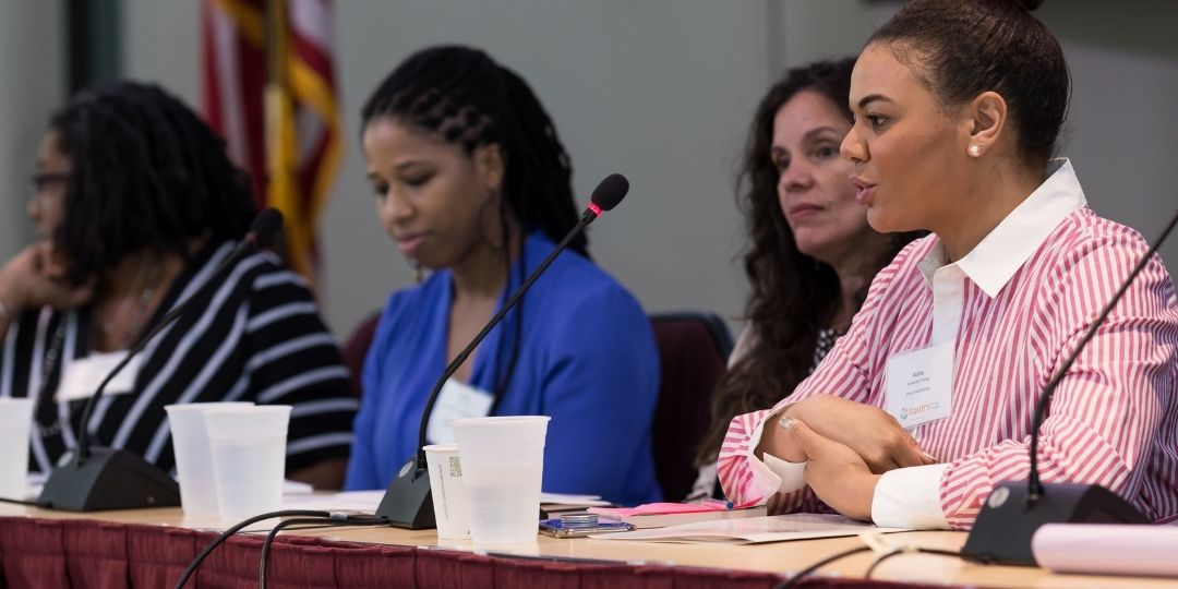 Four people sitting at a presentation table with microphones. Person speaking is wearing a collared pinstripe pink and white shirt.