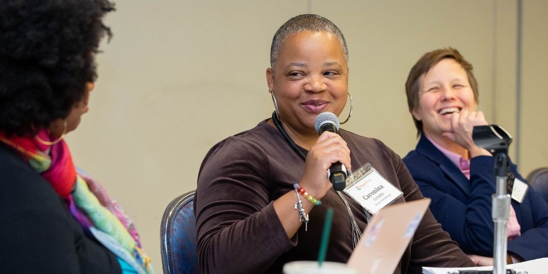 Three people sitting down at conference table presenting. Middle person has a microphone and person behind them is smiling