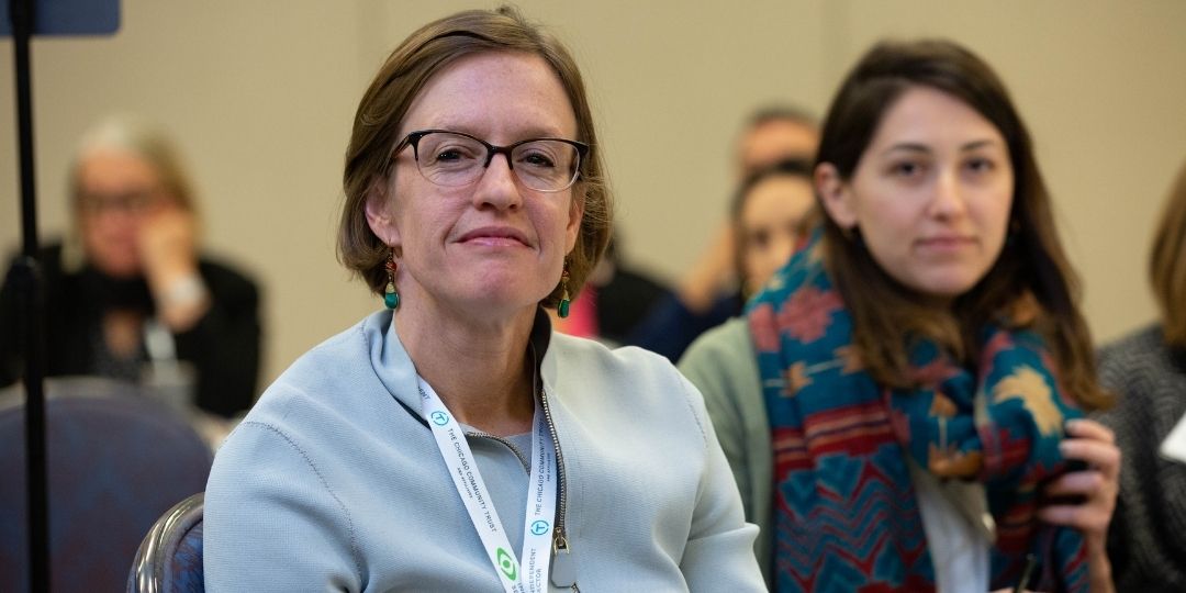 Two people sitting down at conference looking into camera. Other attendees in the background.